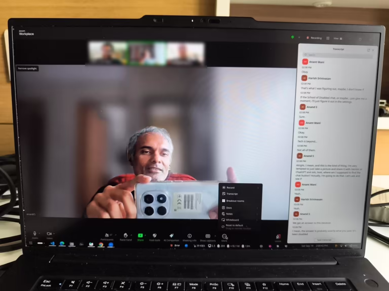 A phone-camera photo of Anand's laptop screen showing the Zoom 'More' menu — Record, Transcript, Breakout rooms, Docs, Notes, Whiteboard — with no Chat option, and a live transcript panel open on the right.