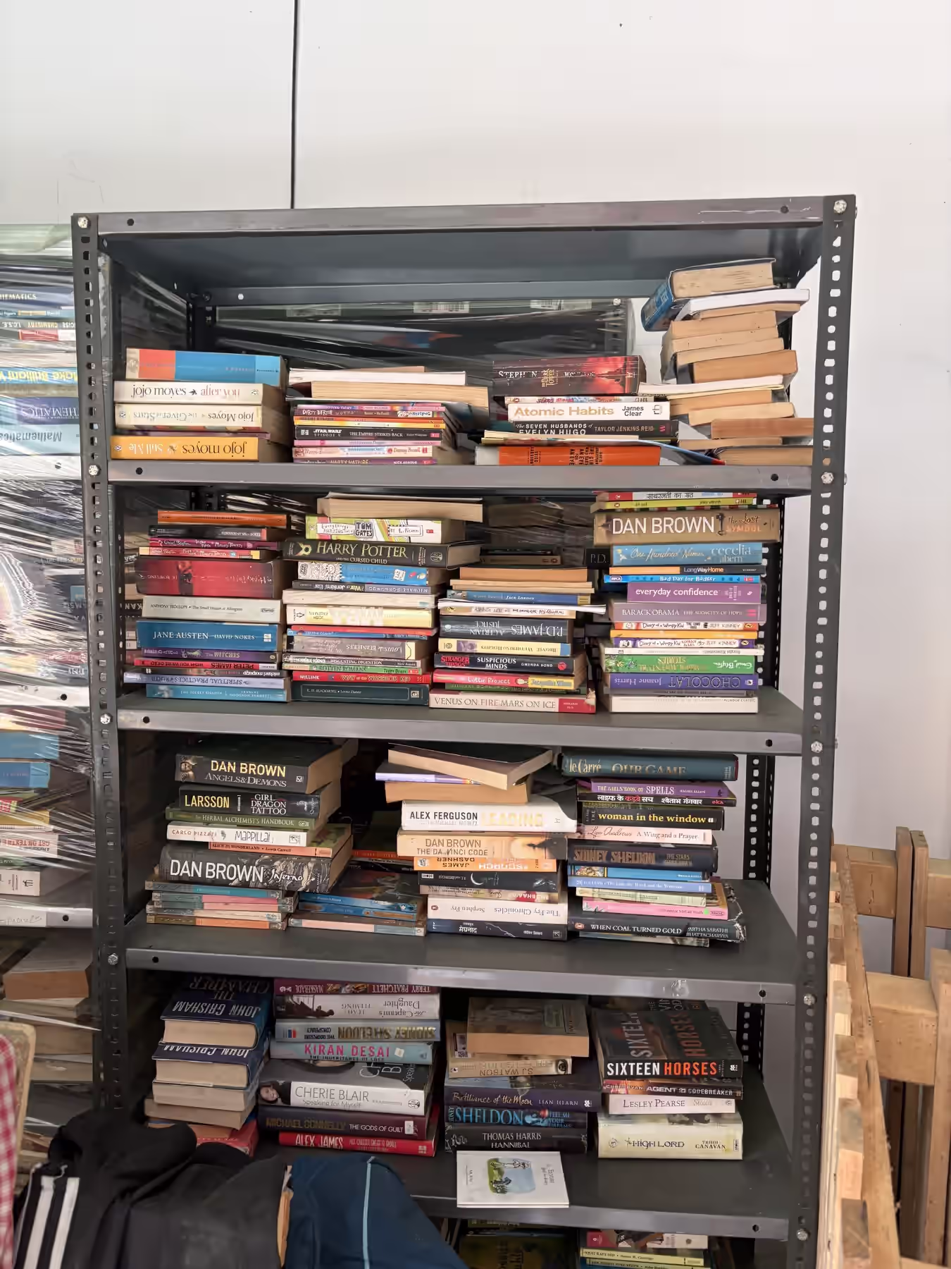 A grey metal rack of stacked paperbacks, spines facing out at many angles: Dan Brown, Jojo Moyes, Sidney Sheldon, Harry Potter and the Cursed Child, Atomic Habits, Alex Ferguson's Leading, and dozens more, some in Hindi, many partly hidden.